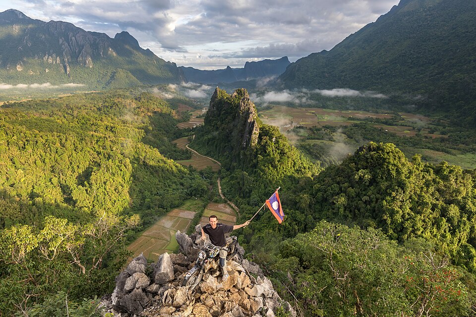 man on a motorcycle holding a flag of laos in front of green karst peaks at the top of mount nam xa