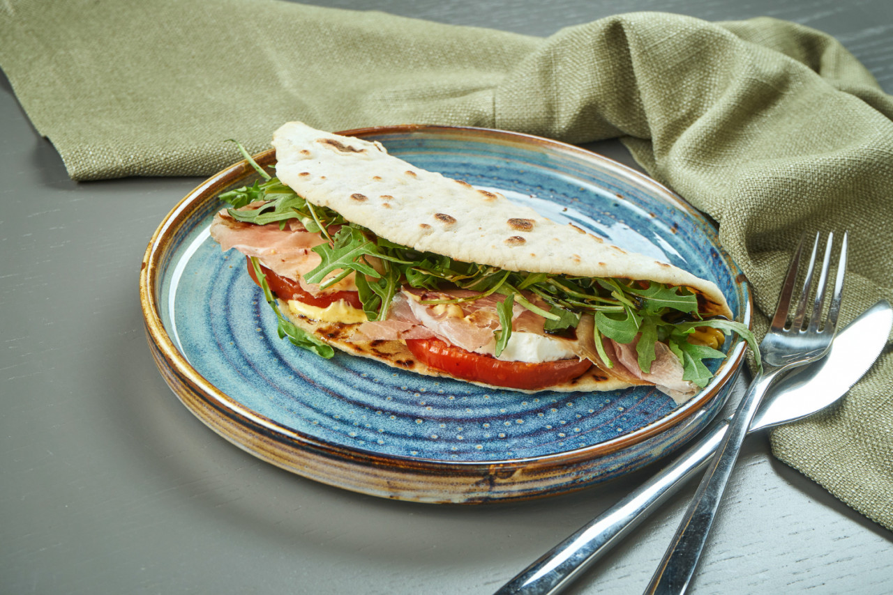 piadina classic italian tortilla bread with prosciutto arugula mozzarella tomatoes blue plate wooden table fast sandwich