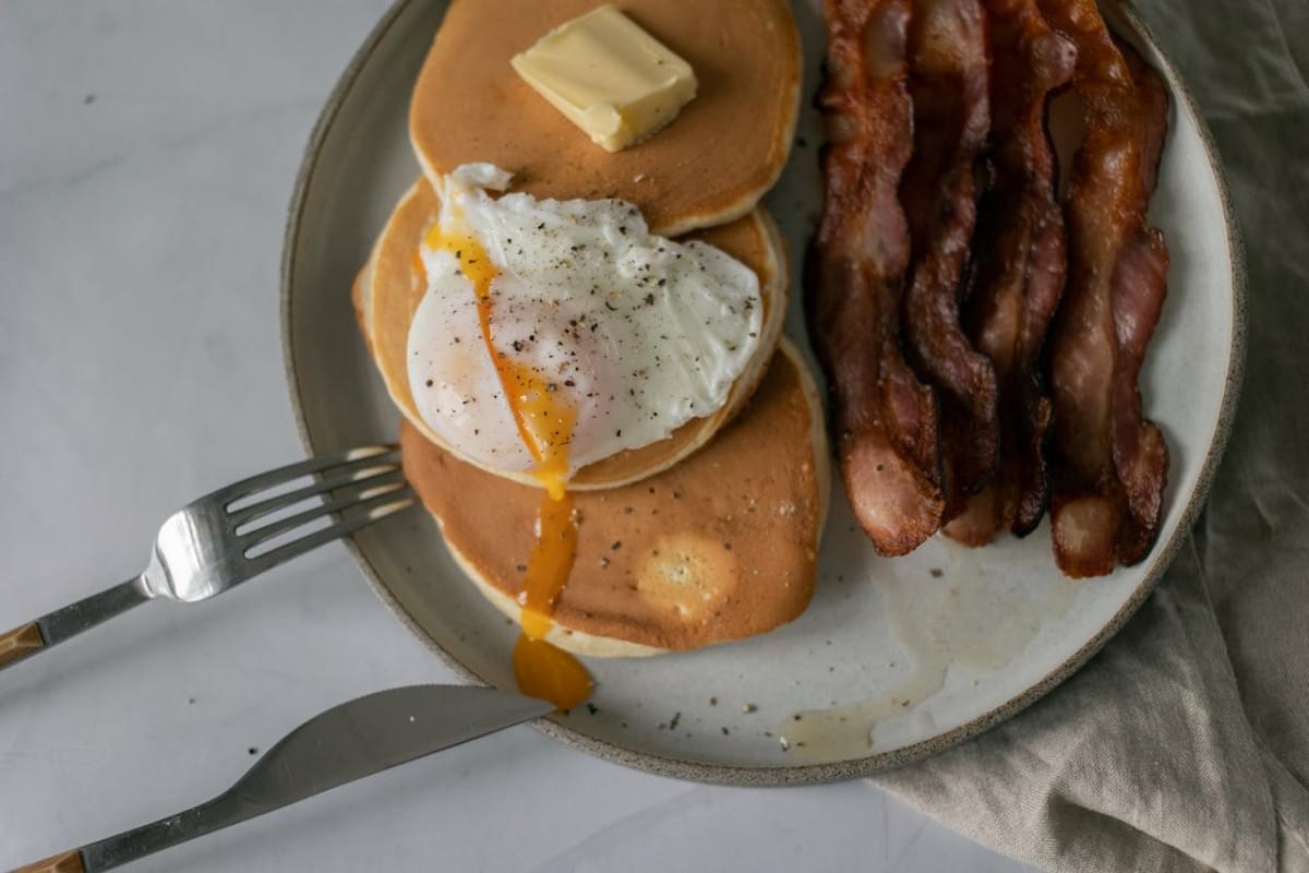 plate with pancakes with butter near poached egg and roasted bacon 1